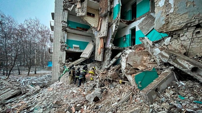 Rescue workers are clearing rubble from a severely damaged residential building. The structure is partially collapsed, with debris scattered around