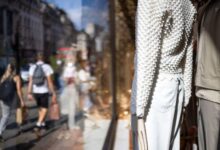 People walk past a clothing store window display in London, featuring mannequins dressed in textured white tops and light-coloured pants. The reflection shows a busy street with shoppers carrying bags.