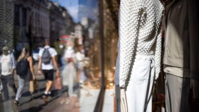 People walk past a clothing store window display in London, featuring mannequins dressed in textured white tops and light-coloured pants. The reflection shows a busy street with shoppers carrying bags.