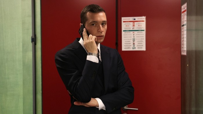 Jordan Bardella is pictured speaking on the phone as he stands in front of a red door